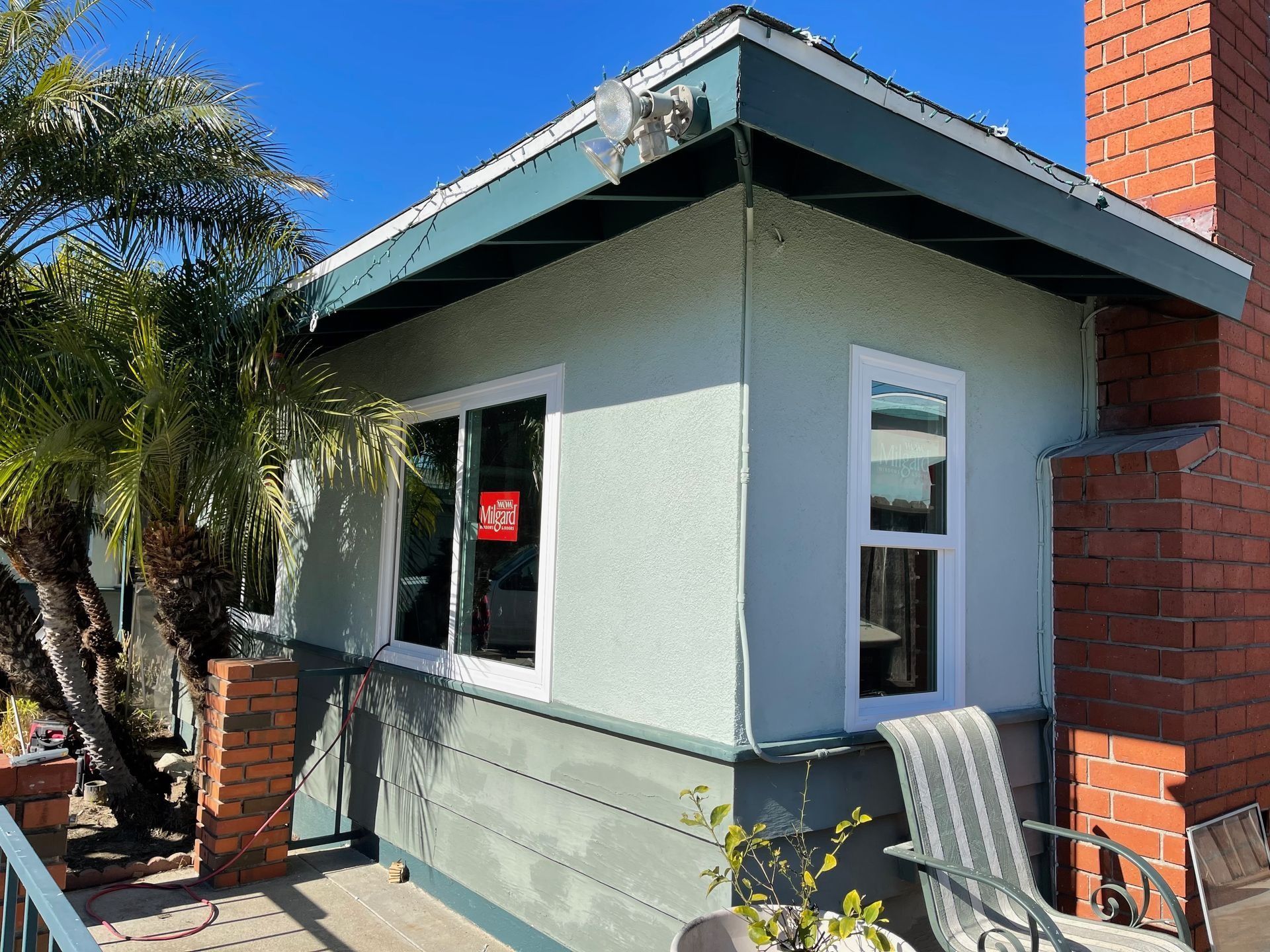 Small building with light green stucco walls and white window frames, next to a brick chimney and palm tree.