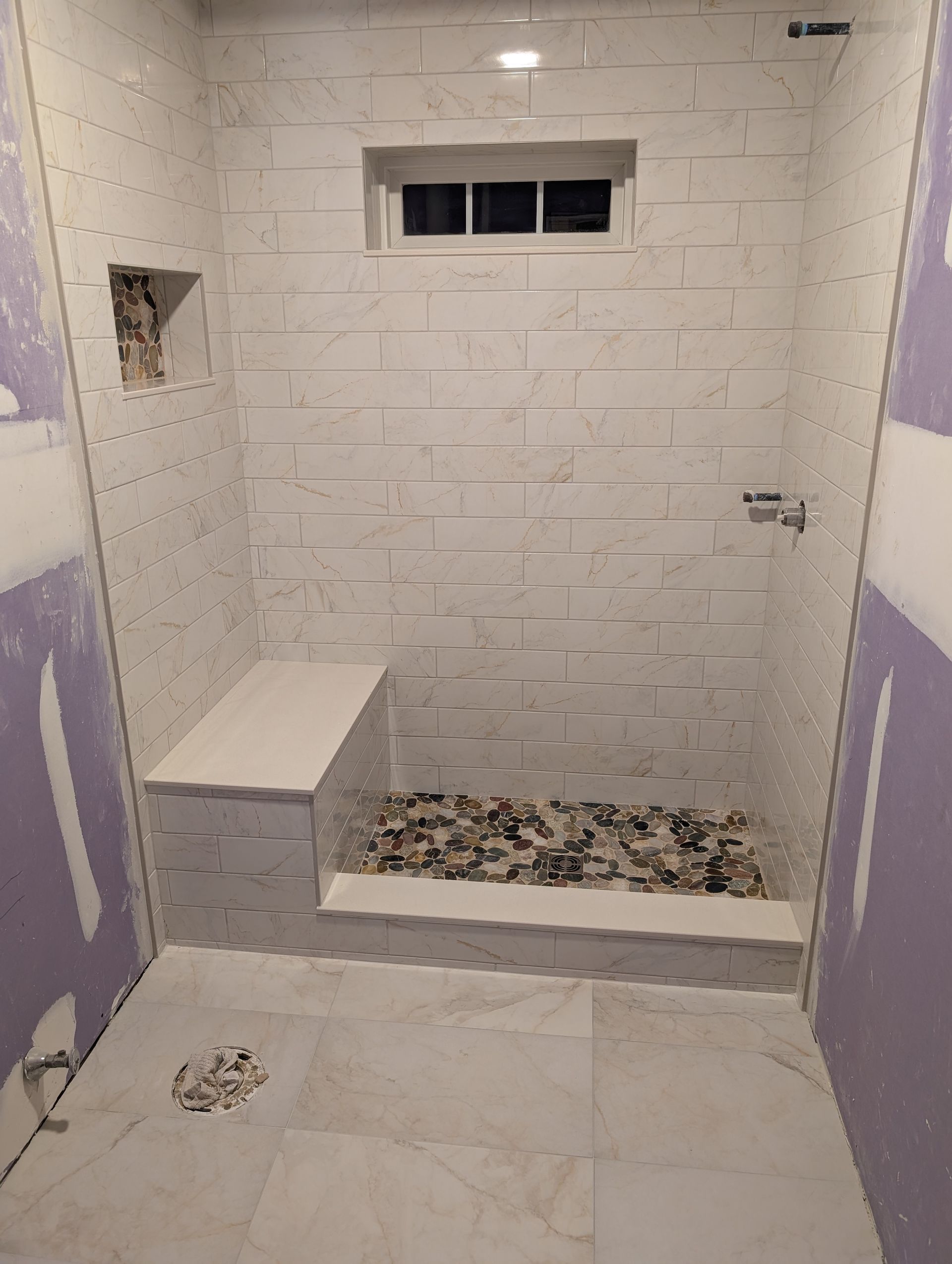 A bathroom shower under construction with white brick-style wall tiles, a stone pebble floor, and a built-in tiled bench.