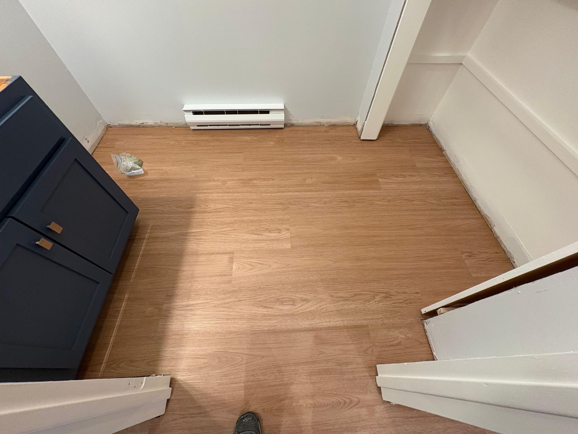 A view of a room with wood-look vinyl flooring, a navy cabinet on the left, and a white baseboard heater on the back wall.