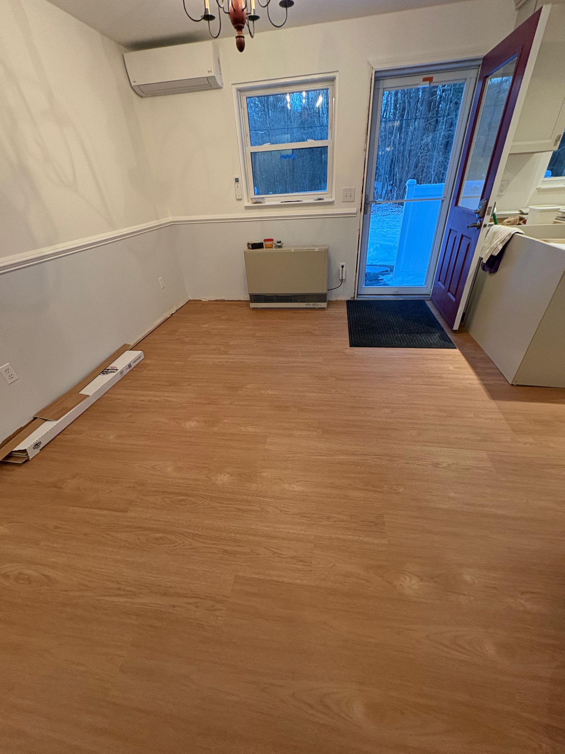 A bright room with new light wood-look flooring, a window, a heater, a white mini-split unit, and an open purple door.