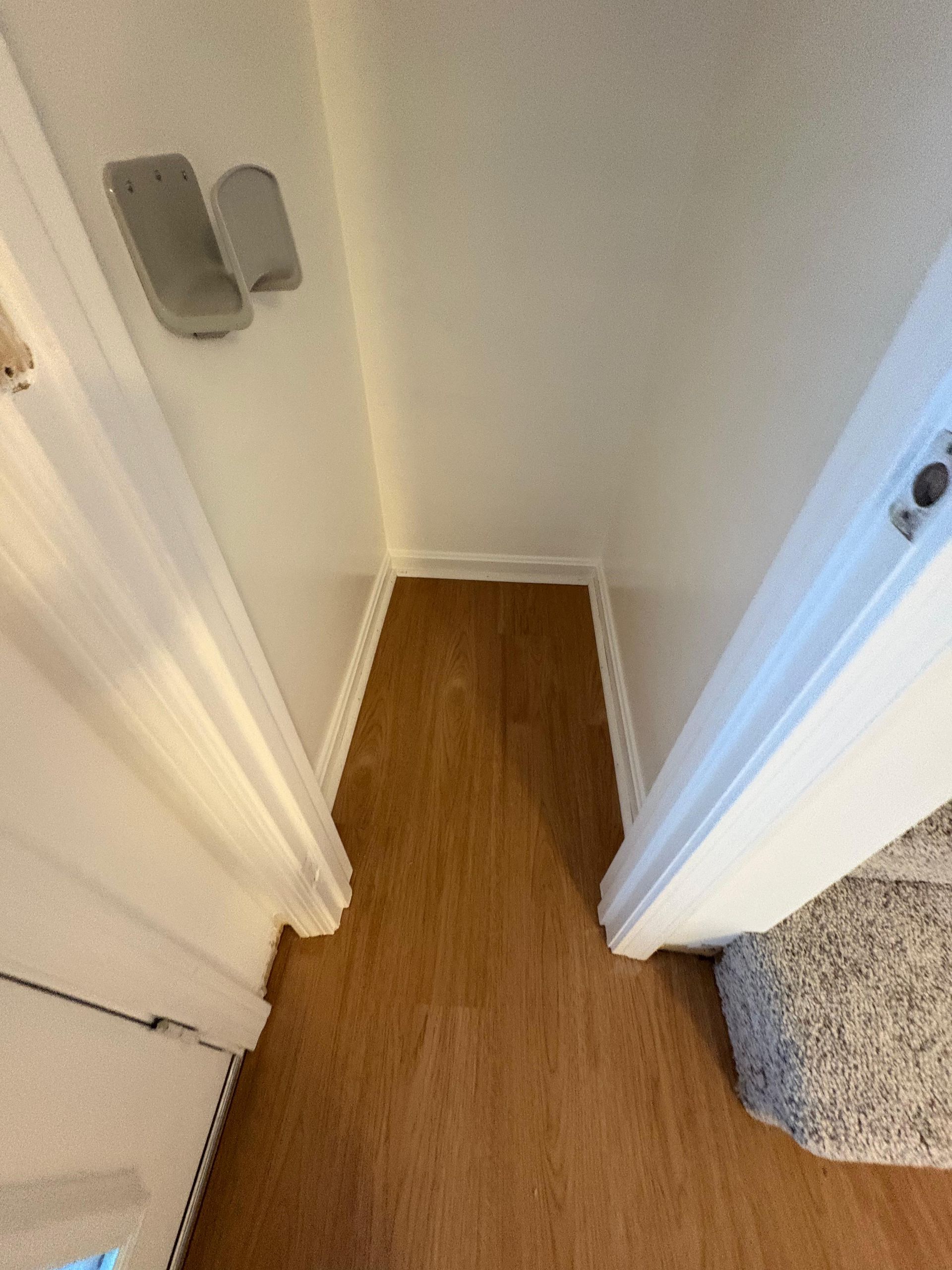 A narrow alcove with light wood flooring and white walls, viewed from a doorway next to a carpeted staircase.