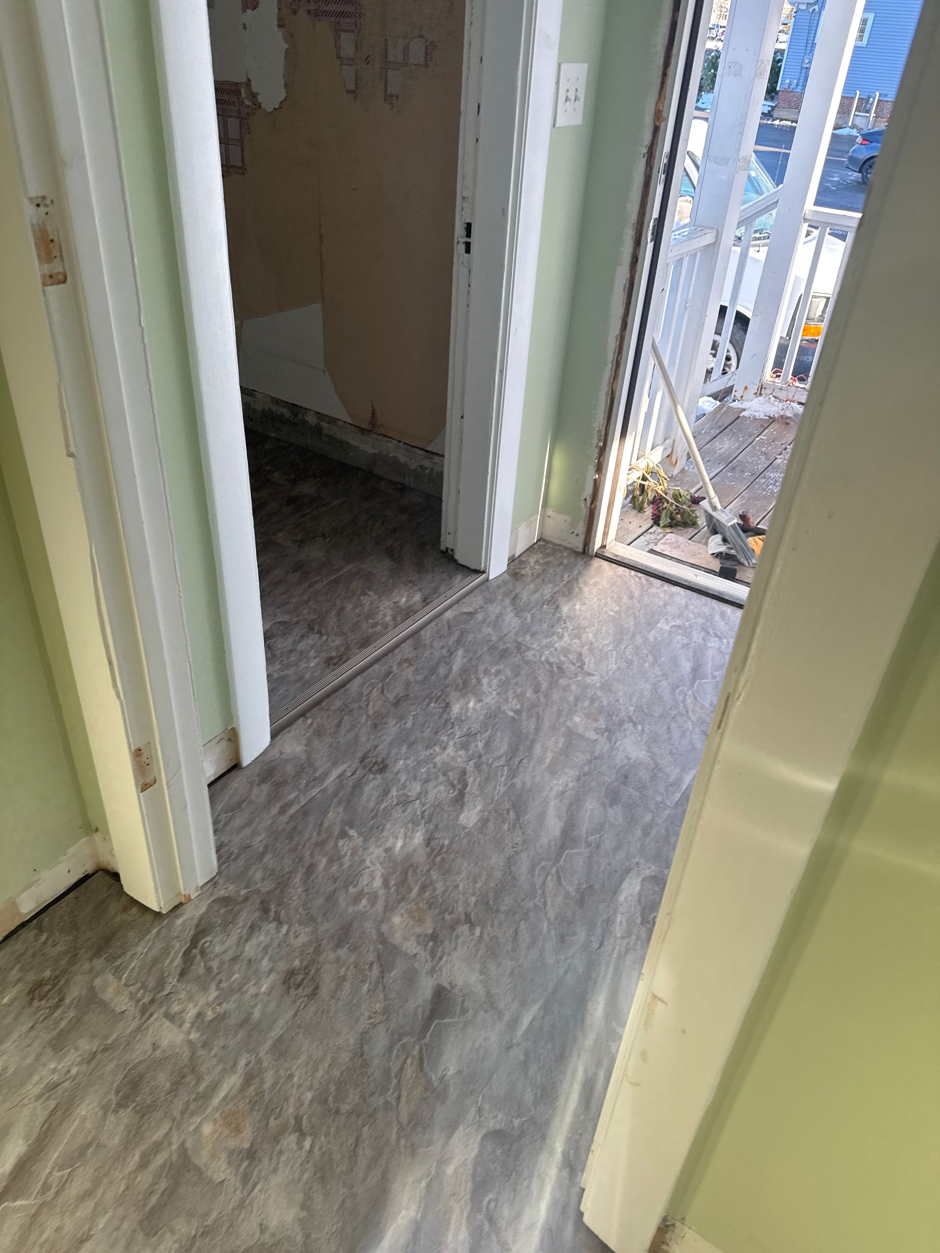 A hallway with grey wood-look flooring leads to an open doorway showing a room with damaged, peeling wallpaper.