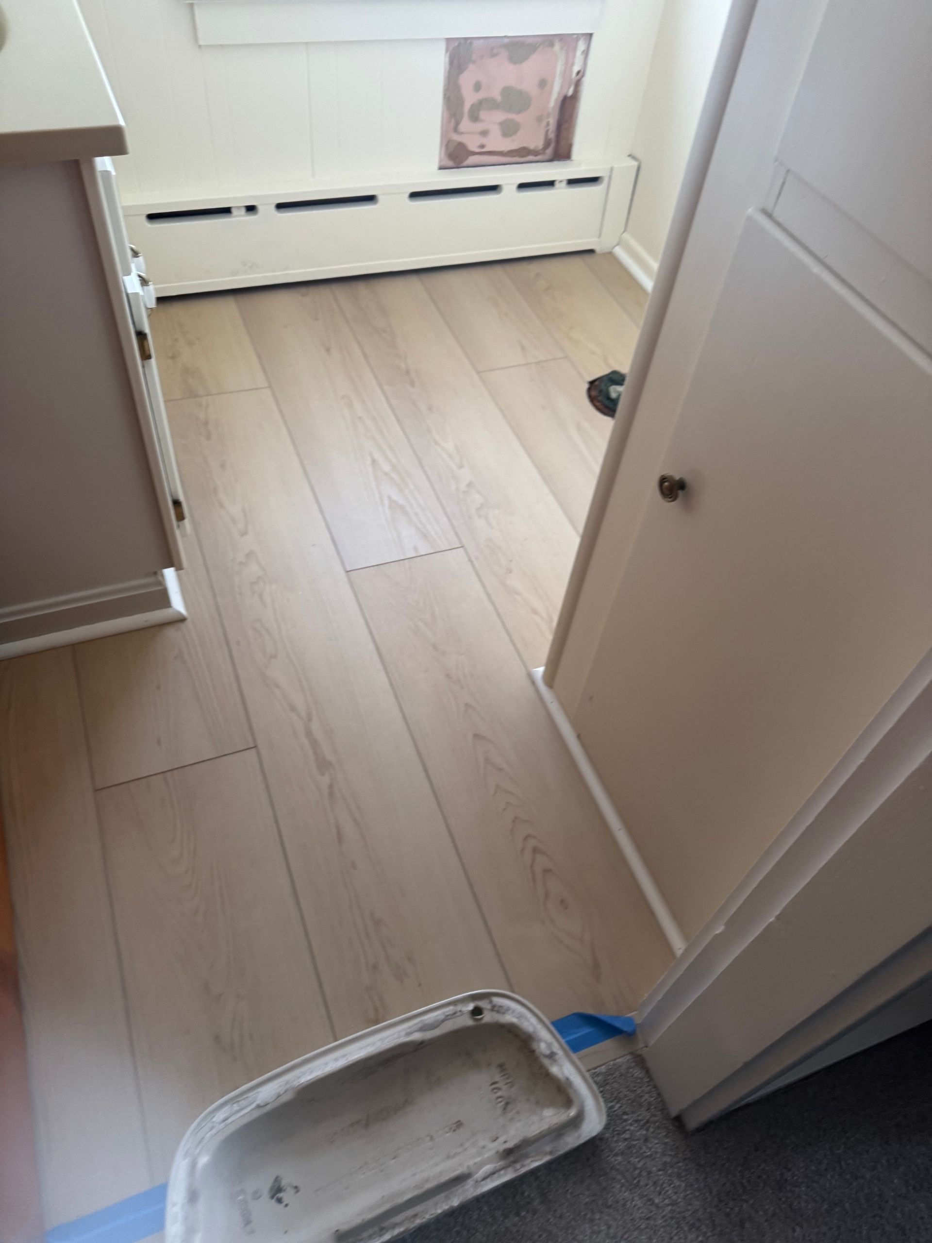 Light-colored wood laminate flooring installed in a room featuring a baseboard heater and a partially open white door.