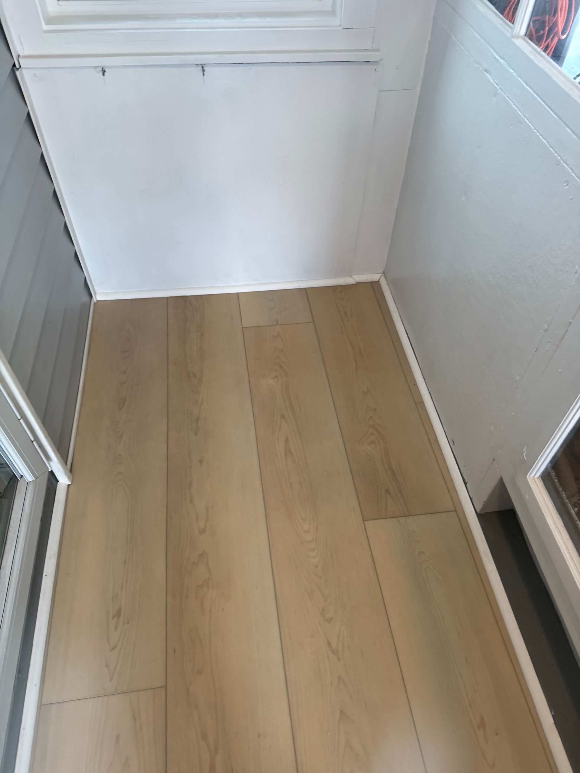 A view looking down at light-toned wood-look laminate flooring installed in a small, enclosed porch area with white walls.