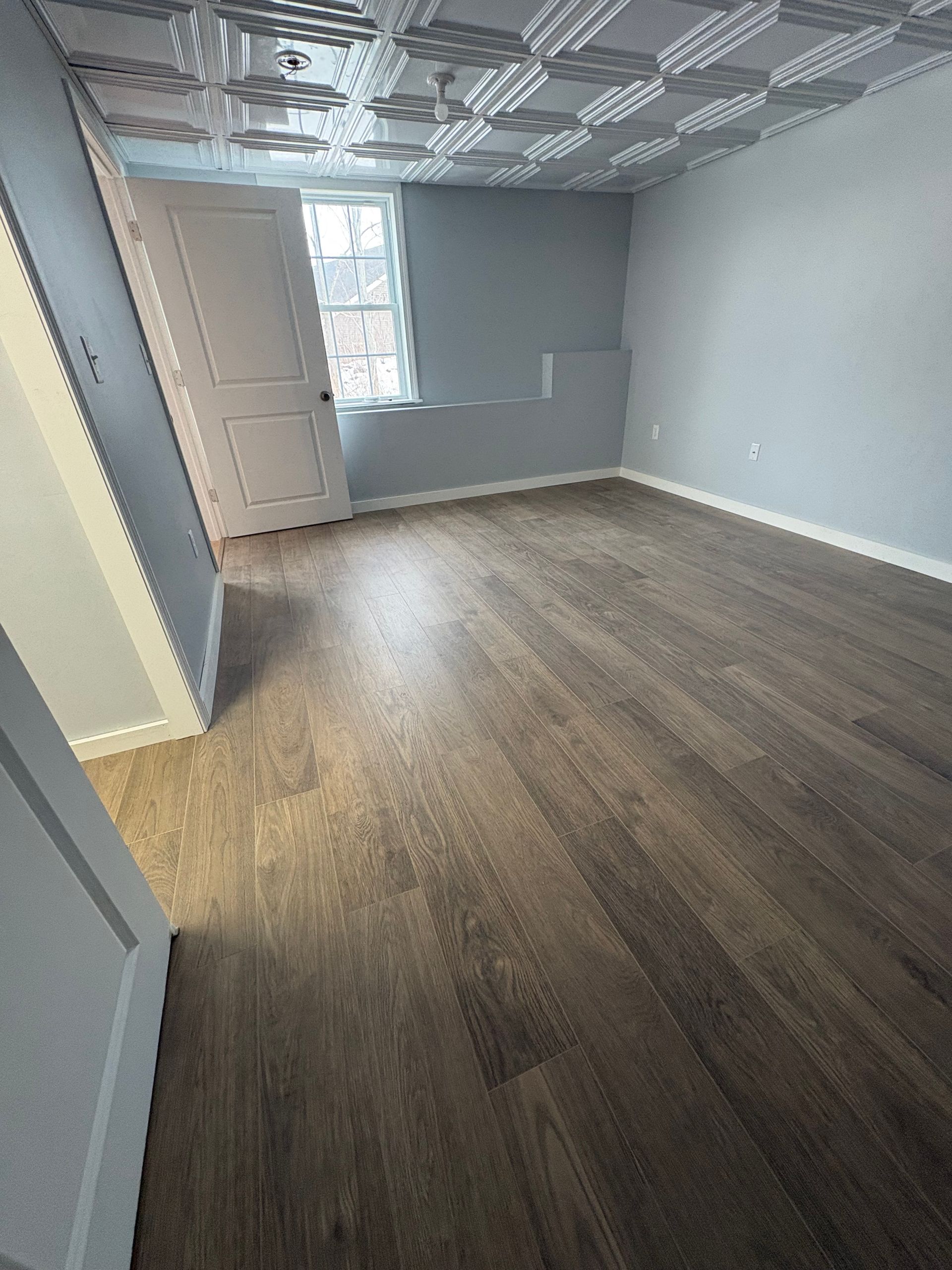An empty room with light gray walls, dark wood-look plank flooring, and a decorative white pressed-tin style ceiling.