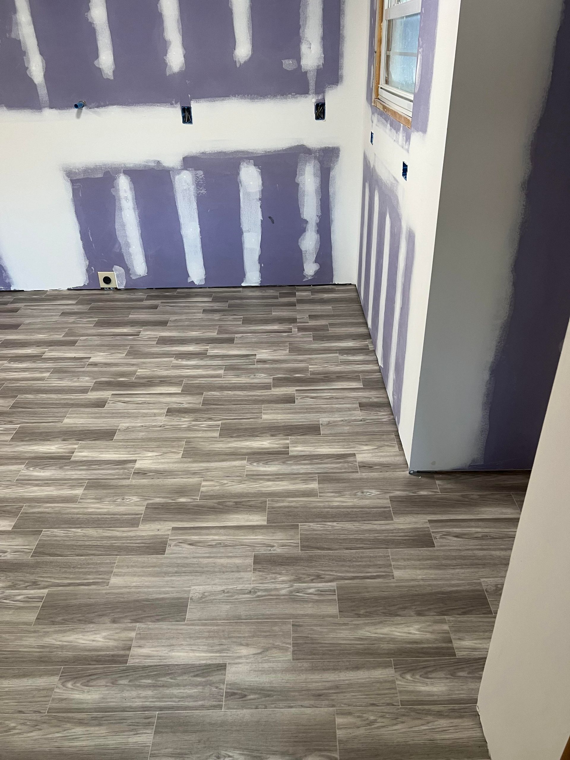 A room under construction with gray wood-look plank flooring and drywall panels that have been taped and mudded.