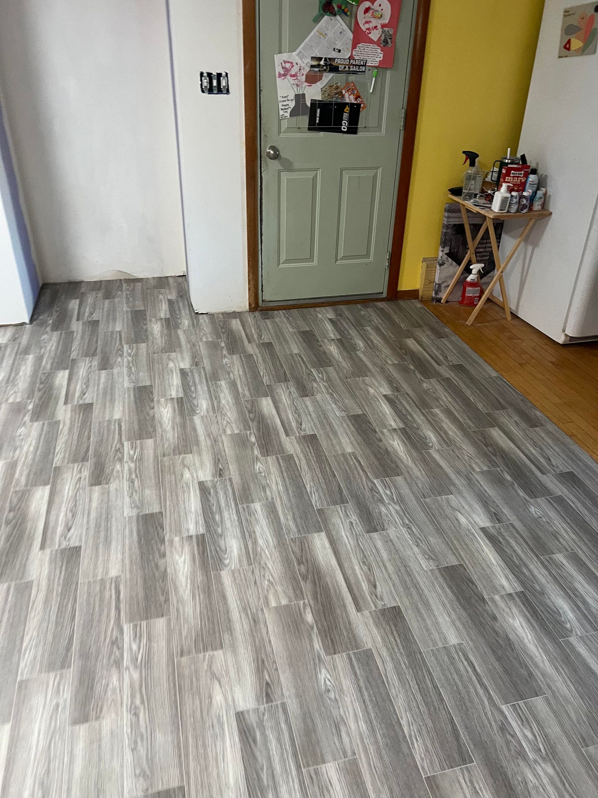 Newly installed gray wood-look laminate flooring in a room next to a green door and a yellow wall with a small side table.