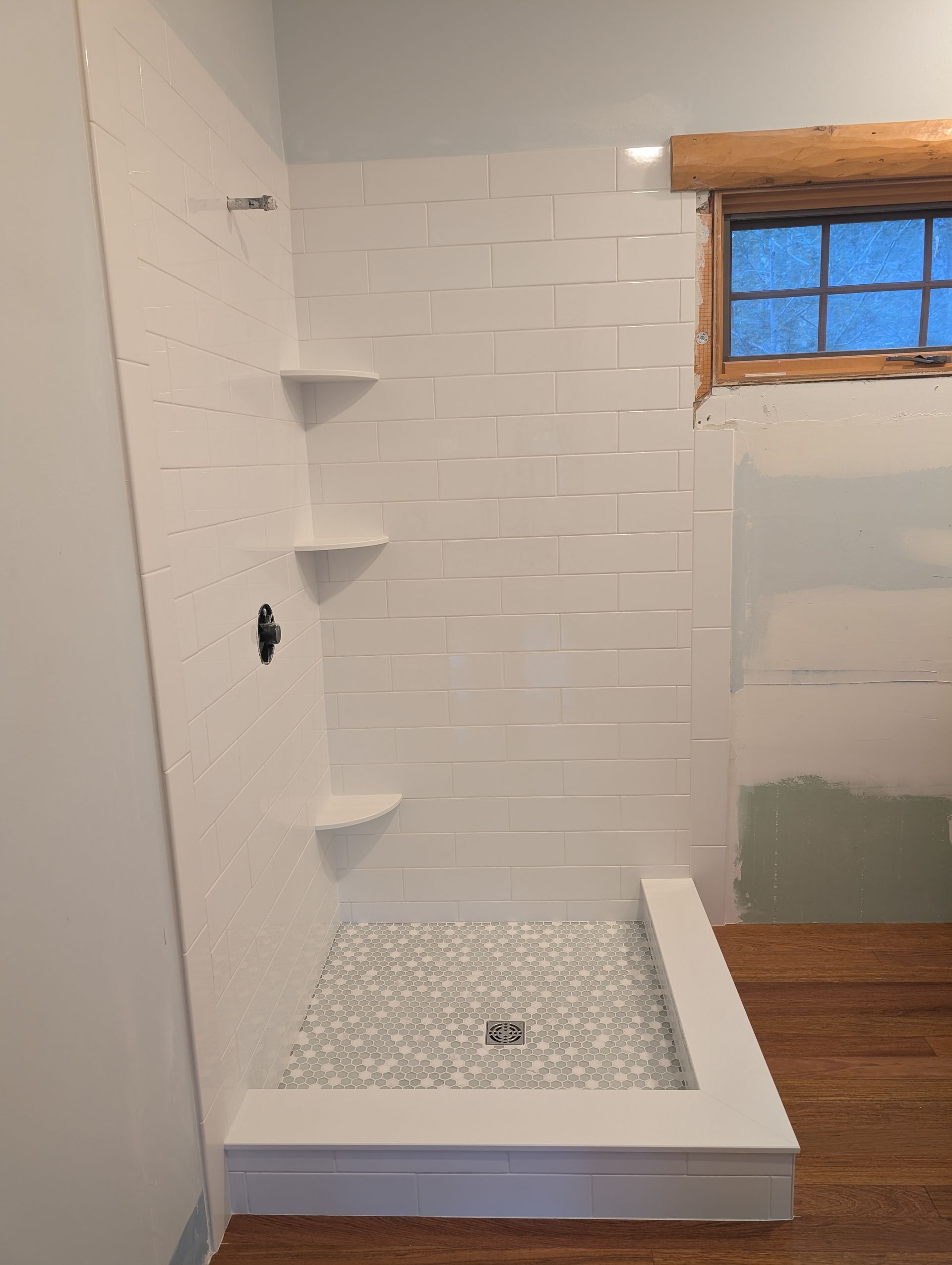 A tiled shower stall with white brick-style walls, three corner shelves, a gray pebble-patterned floor, and a wooden window.