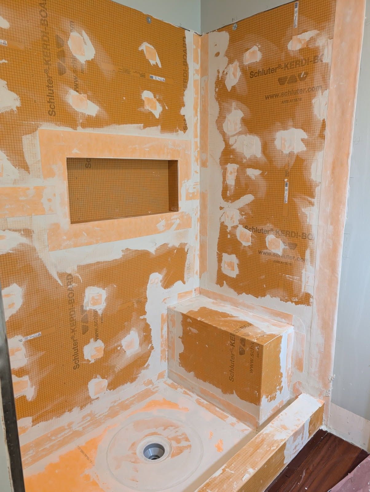 Shower stall under construction with orange waterproof membrane on walls, a niche, a built-in bench, and a drain.