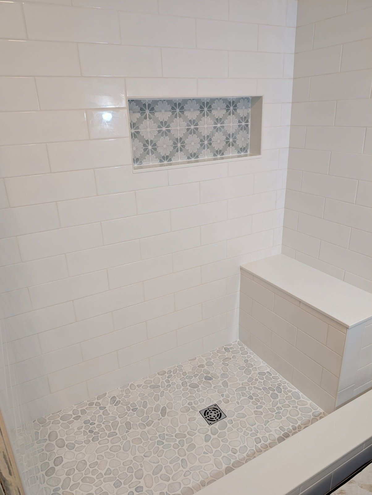 A walk-in shower with white subway tile walls, a decorative patterned tile niche, and a light-colored stone pebble floor.