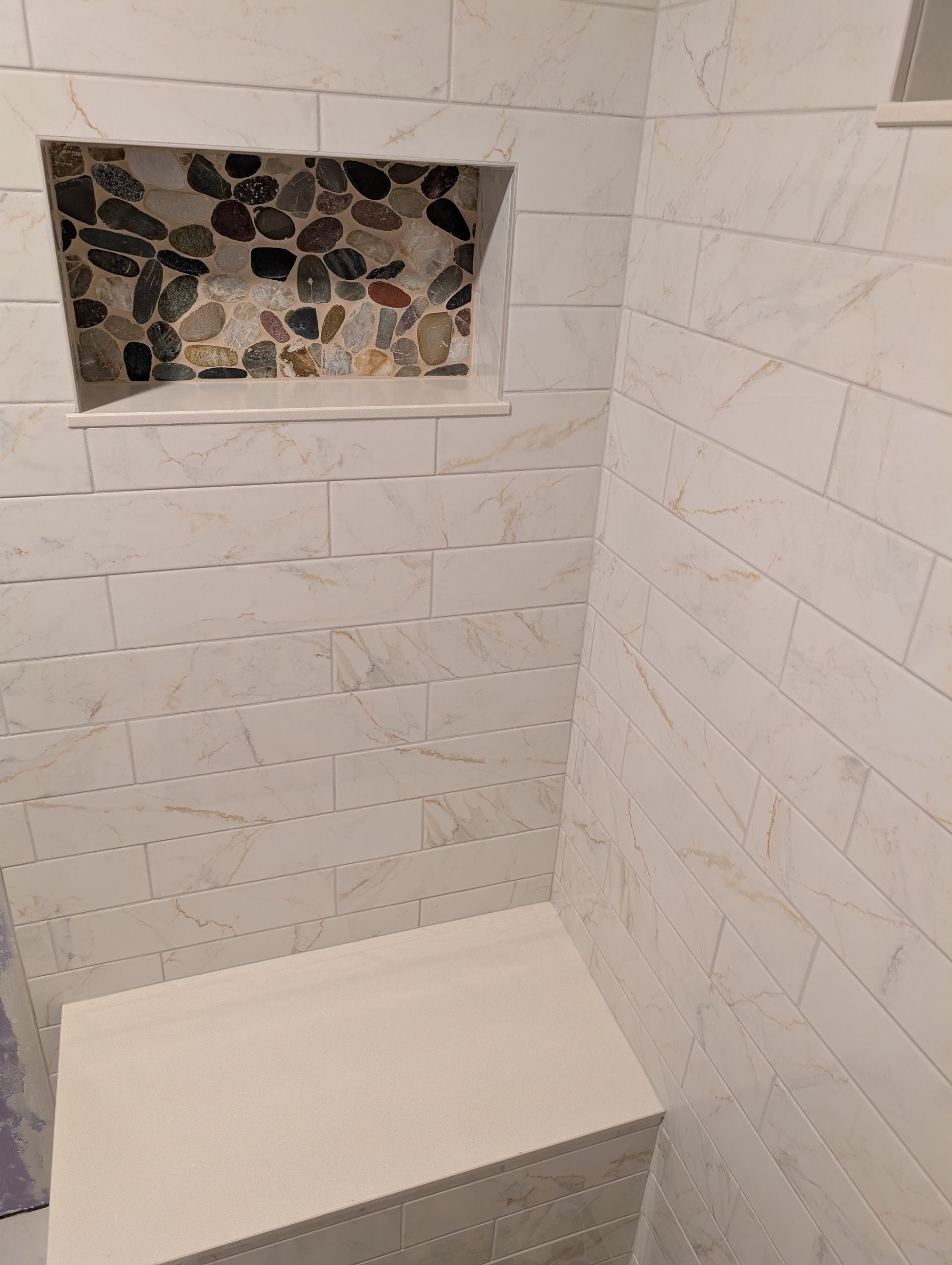 A shower interior featuring white marble-look subway tiles, a built-in stone bench, and a niche with decorative pebble tile.