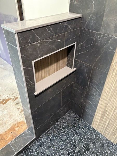 Tiled shower corner with dark gray walls, built-in shelf, and pebble floor. Light wood-look tile on the right.