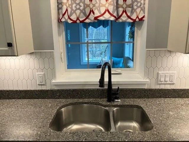Kitchen sink with a black faucet, surrounded by granite countertop and white backsplash tile. A window is centered above.