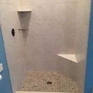 Tiled shower with a triangular seat and shelves. Beige walls, mosaic floor. Black shower fixture.