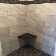 Corner shower with gray tile walls, black pebble floor, and a dark corner shelf.