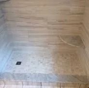 Shower with textured stone tile, gray and tan tones, with a drain in the corner.