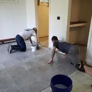 Two people on hands and knees, wiping a concrete floor in a room with doorways.