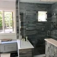 Modern bathroom with gray tile, glass shower, window, vanity with a light countertop.