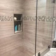 Shower with beige tiled walls, a mosaic border, built-in shelf with toiletries, and glass door.