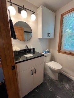 Bathroom with white vanity, black countertop, round mirror, and a wall-mounted cabinet. A toilet sits nearby, and there is a window.
