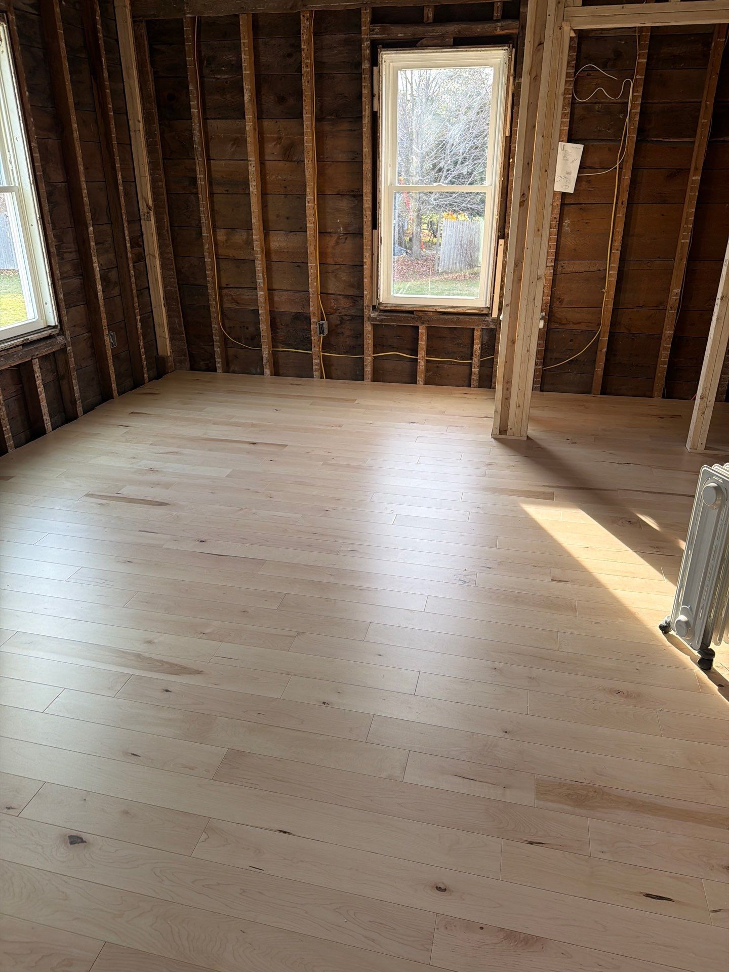 Empty room with newly installed, light-colored hardwood floor. Exposed wood framing and window.