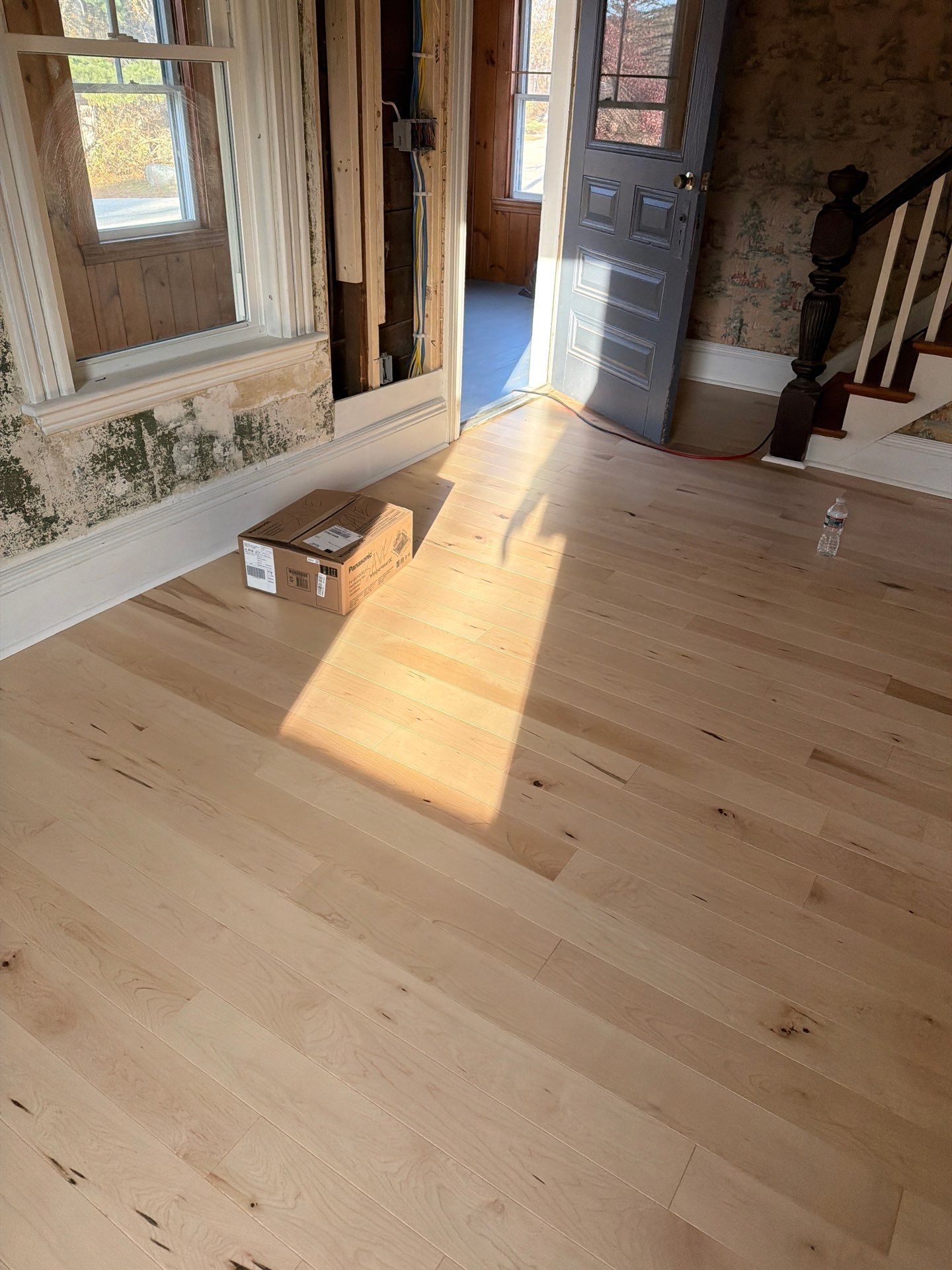Newly installed light wood flooring in a room with a box and partially visible walls. Sunlight illuminates the floor.