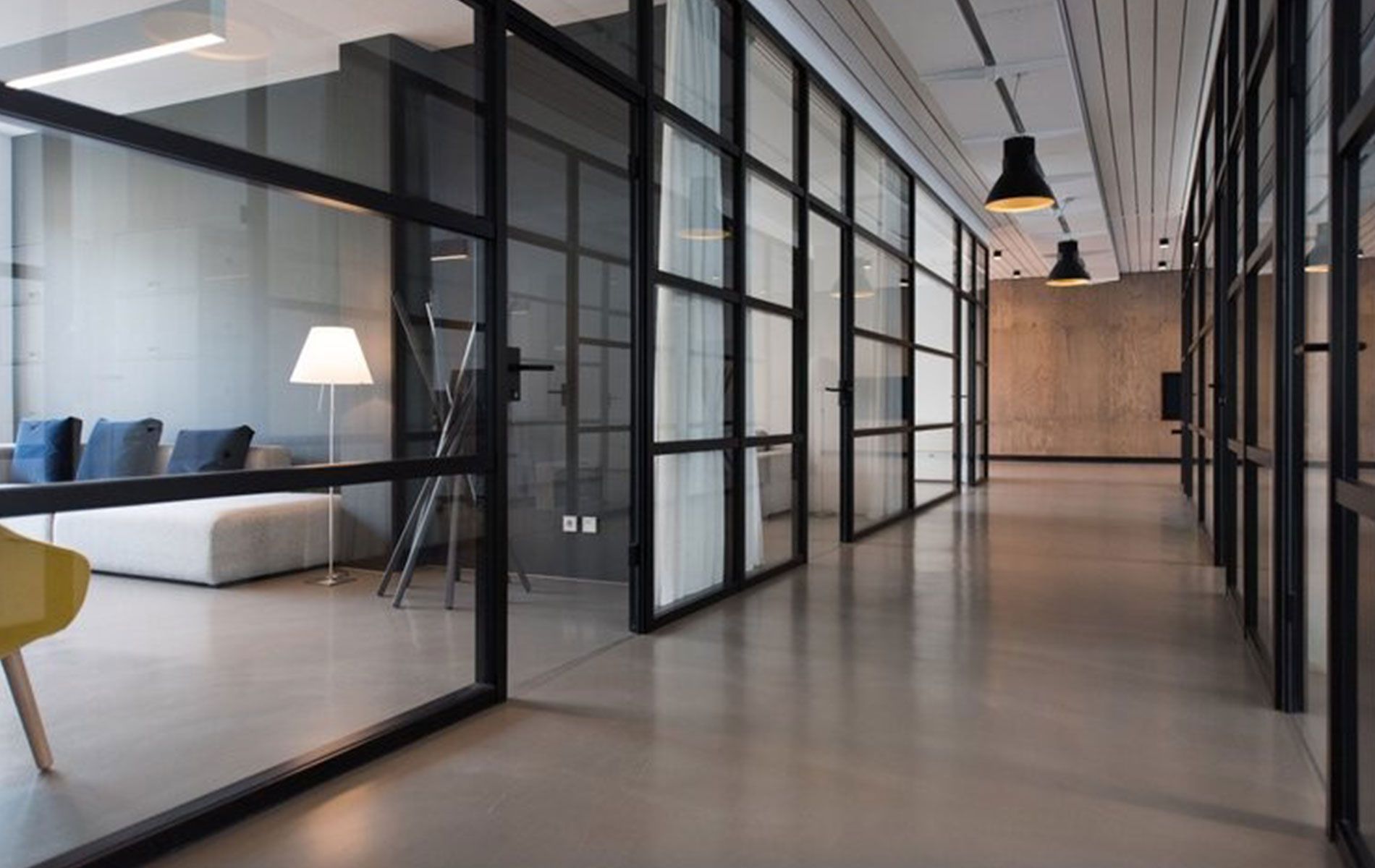 Modern office hallway with glass-walled rooms, gray walls, and pendant lights. A sofa and chair are visible.