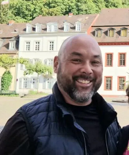 Man with dark hair and beard smiles in front of historic buildings. He wears a black shirt and blue vest.
