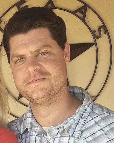 Man with dark hair and blue eyes, smiling, wearing a plaid shirt, in front of a metal Texas sign.