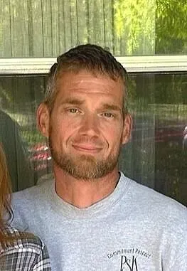 Man with short hair and beard wearing gray shirt, smiling in front of window.