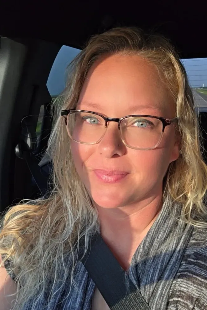 Woman with blonde hair and glasses smiles in a car, sunlit from the side.