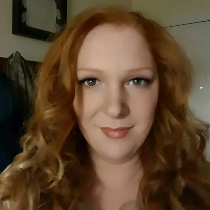 Woman with red wavy hair smiles at the camera, wearing makeup.