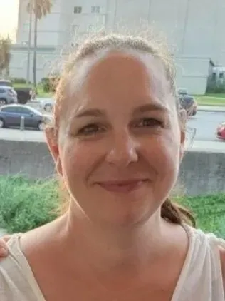Woman smiling, outdoors, neutral expression. Hair pulled back. Buildings and cars in background.