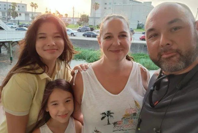 Family of four smiles for a photo outdoors. Buildings, cars, and the sky are in the background.