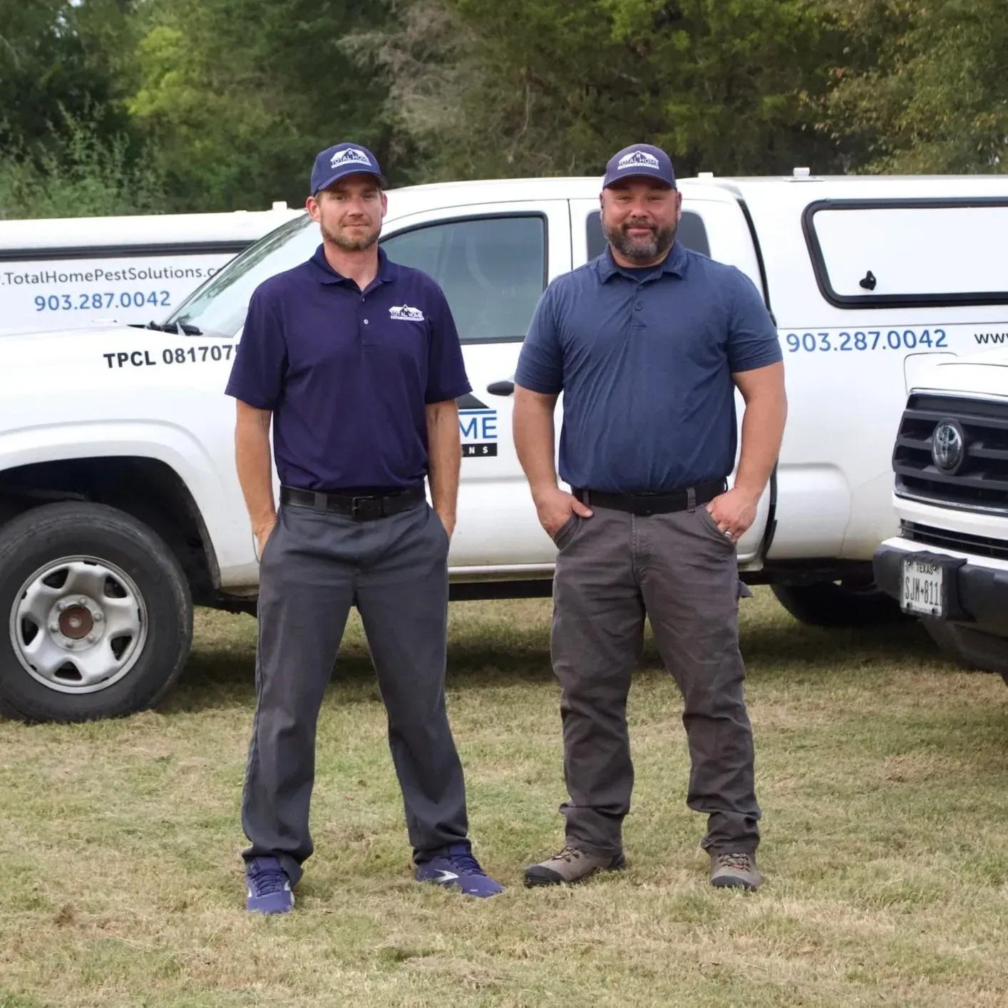 Two men in matching shirts and hats stand in front of white trucks, smiling.