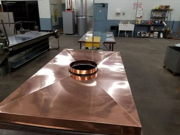 Copper range hood in a workshop, with a round vent and reflecting overhead lights.