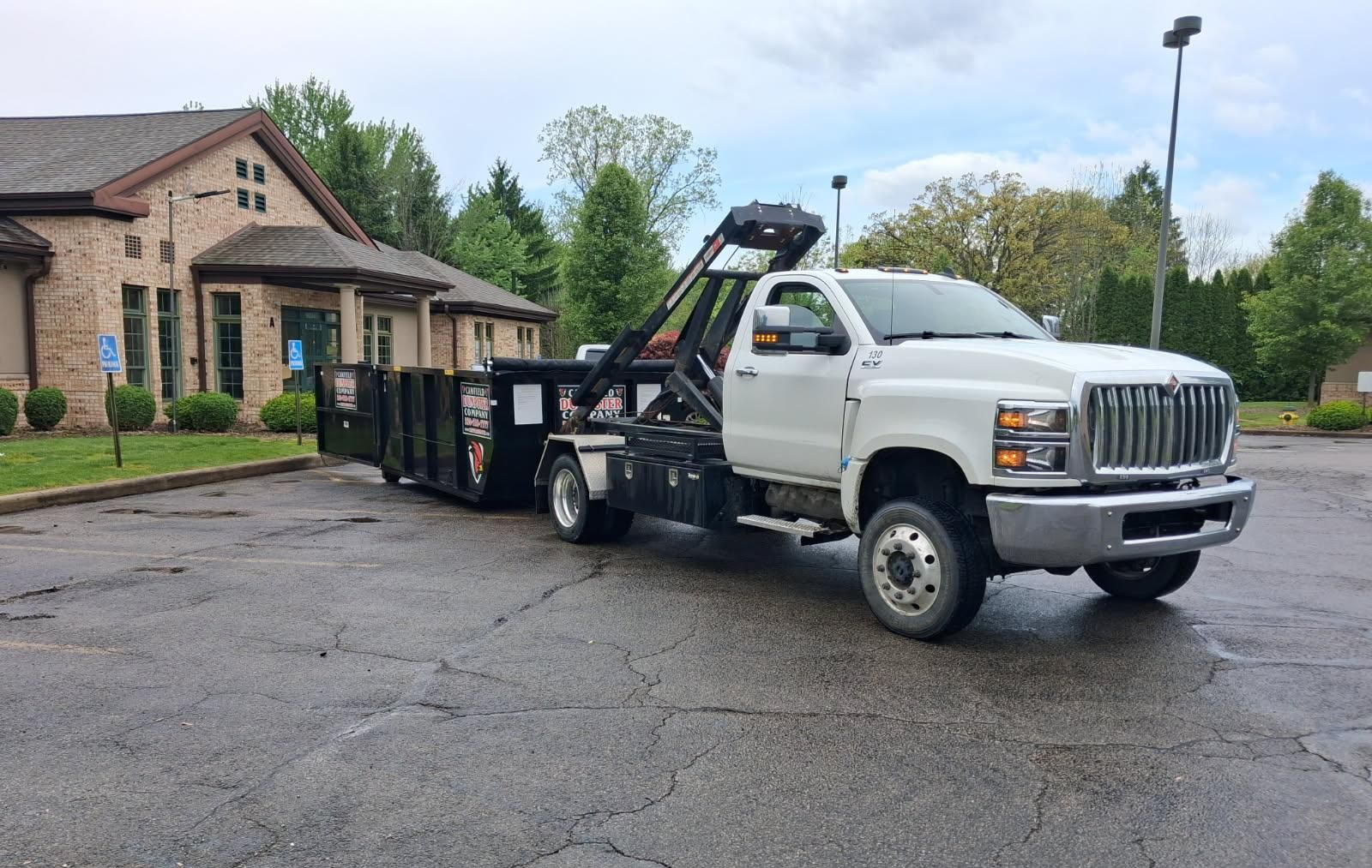 White tow truck loading a dumpster in a parking lot beside a brick house