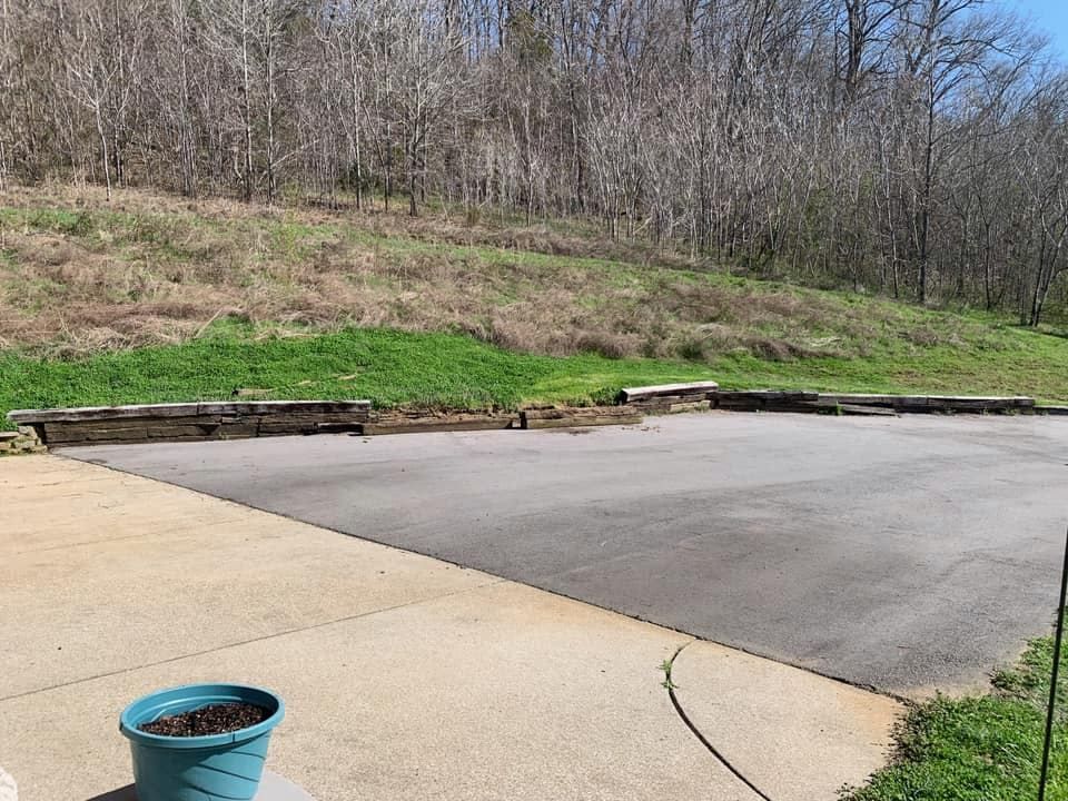 Asphalt parking area bordered by wooden retaining walls and a grassy hillside, with trees in the background and a blue planter in the foreground.