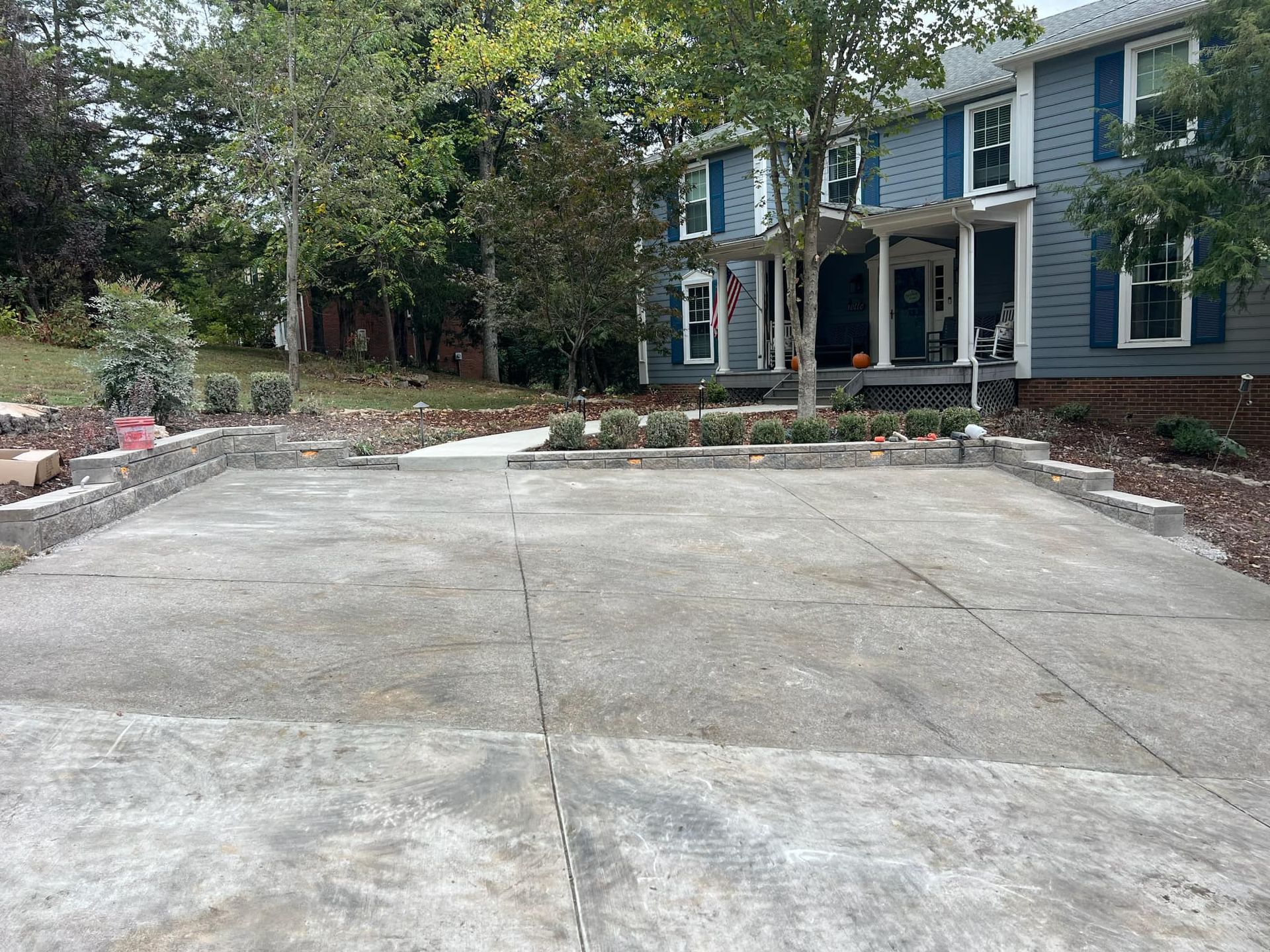 Concrete driveway with stone retaining walls and landscaping in front of a two-story blue house with white columns.