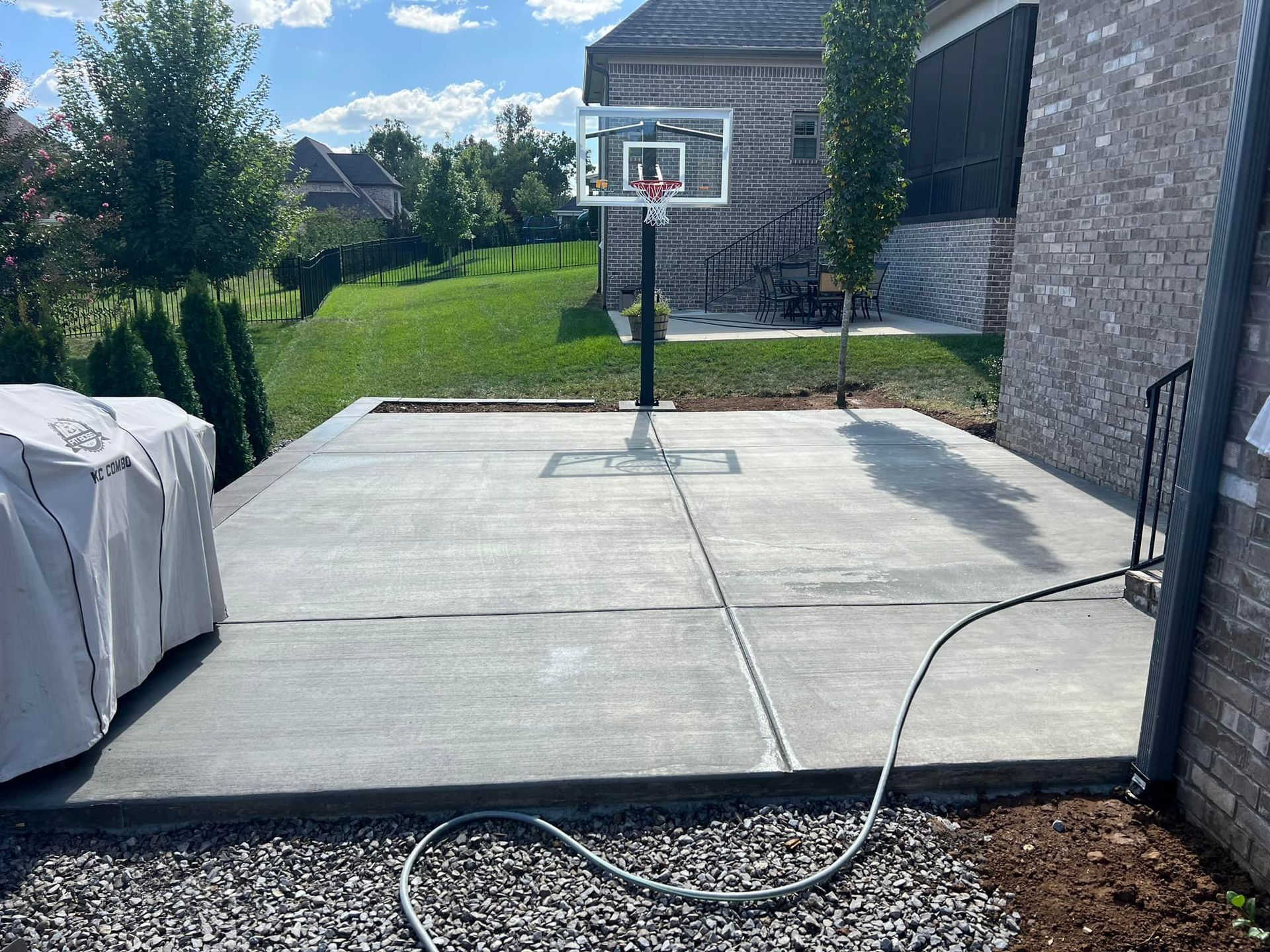 Concrete patio with a basketball hoop, grill, and a grassy hill in the background. A black hose runs across the patio.
