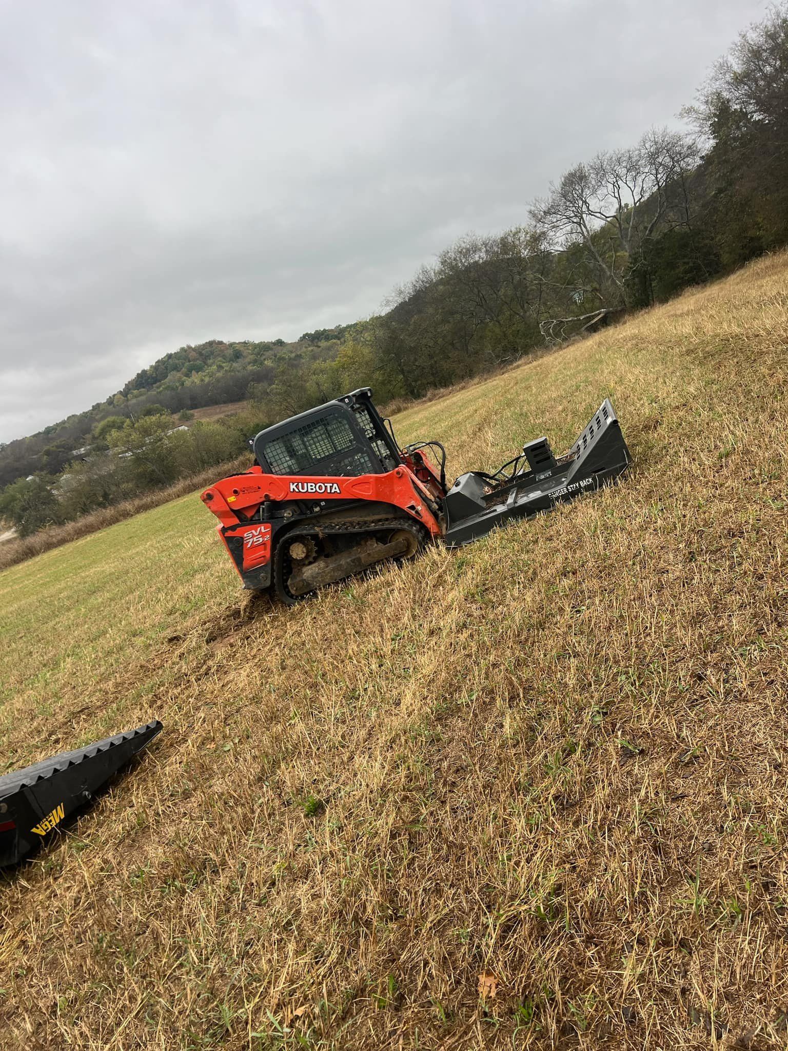 An orange skid steer with a mulching head on a hillside with dry grass. Trees and a cloudy sky are in the background.