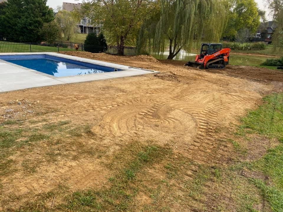 A small orange excavator works near a pool and a lake. The yard shows signs of recent construction.