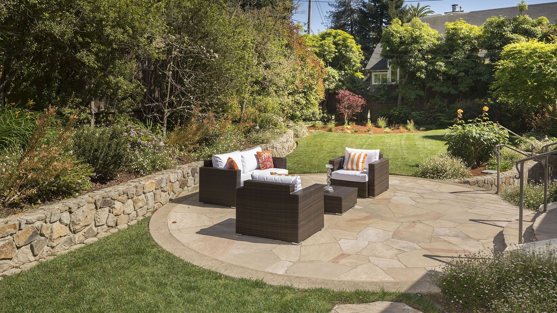 Outdoor patio with wicker furniture on stone, surrounded by grass and a stone retaining wall, trees in the background.