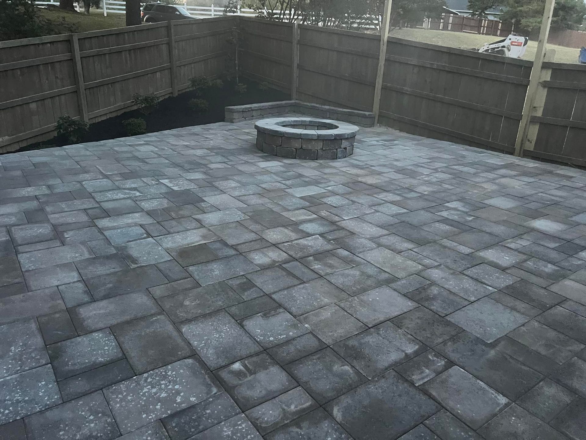 A paved patio with a stone fire pit, enclosed by a wooden fence. The pavers are gray.