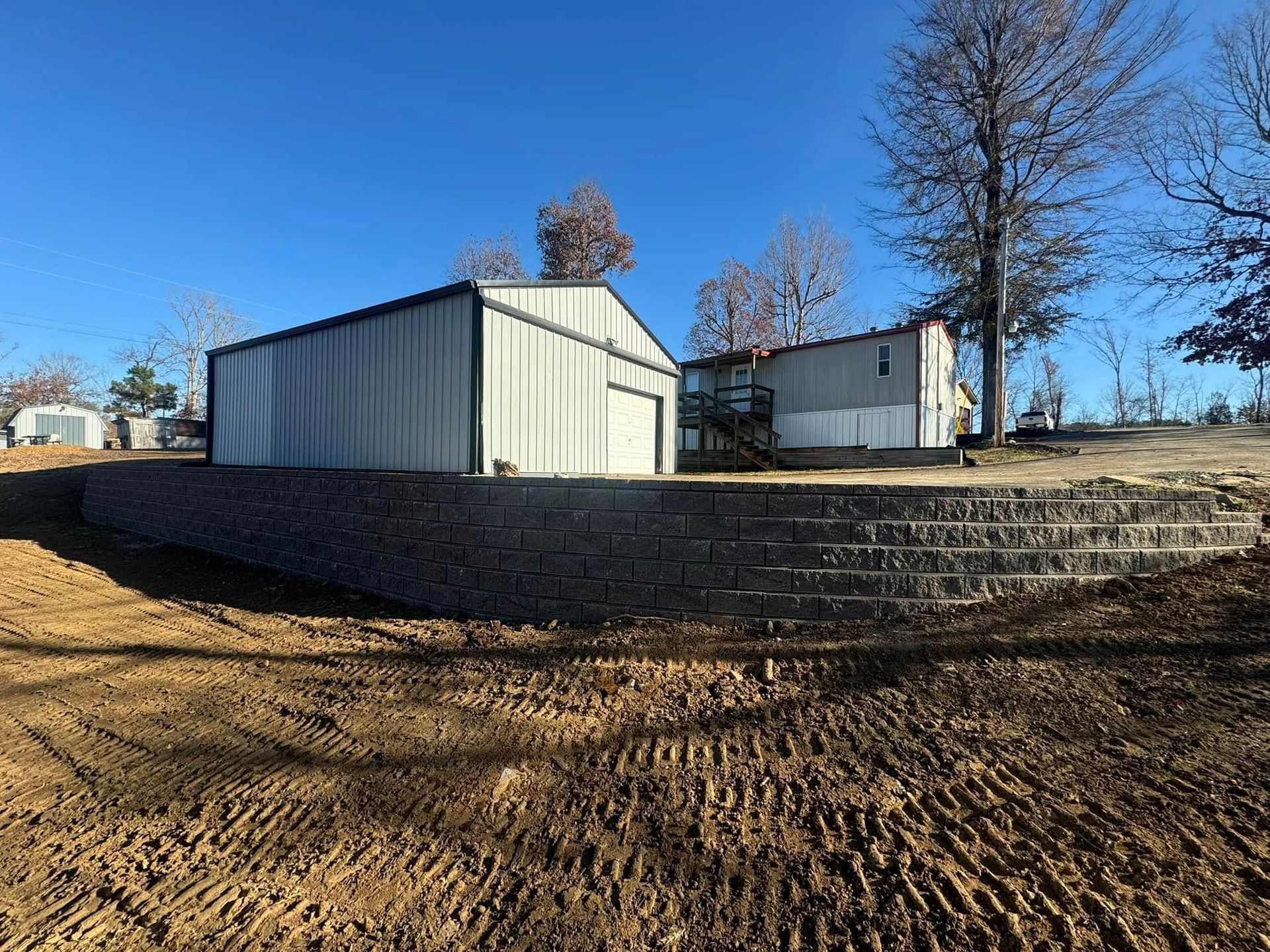 A metal storage building and a mobile home sit behind a stone retaining wall on a sunny day. The foreground is freshly tilled soil.