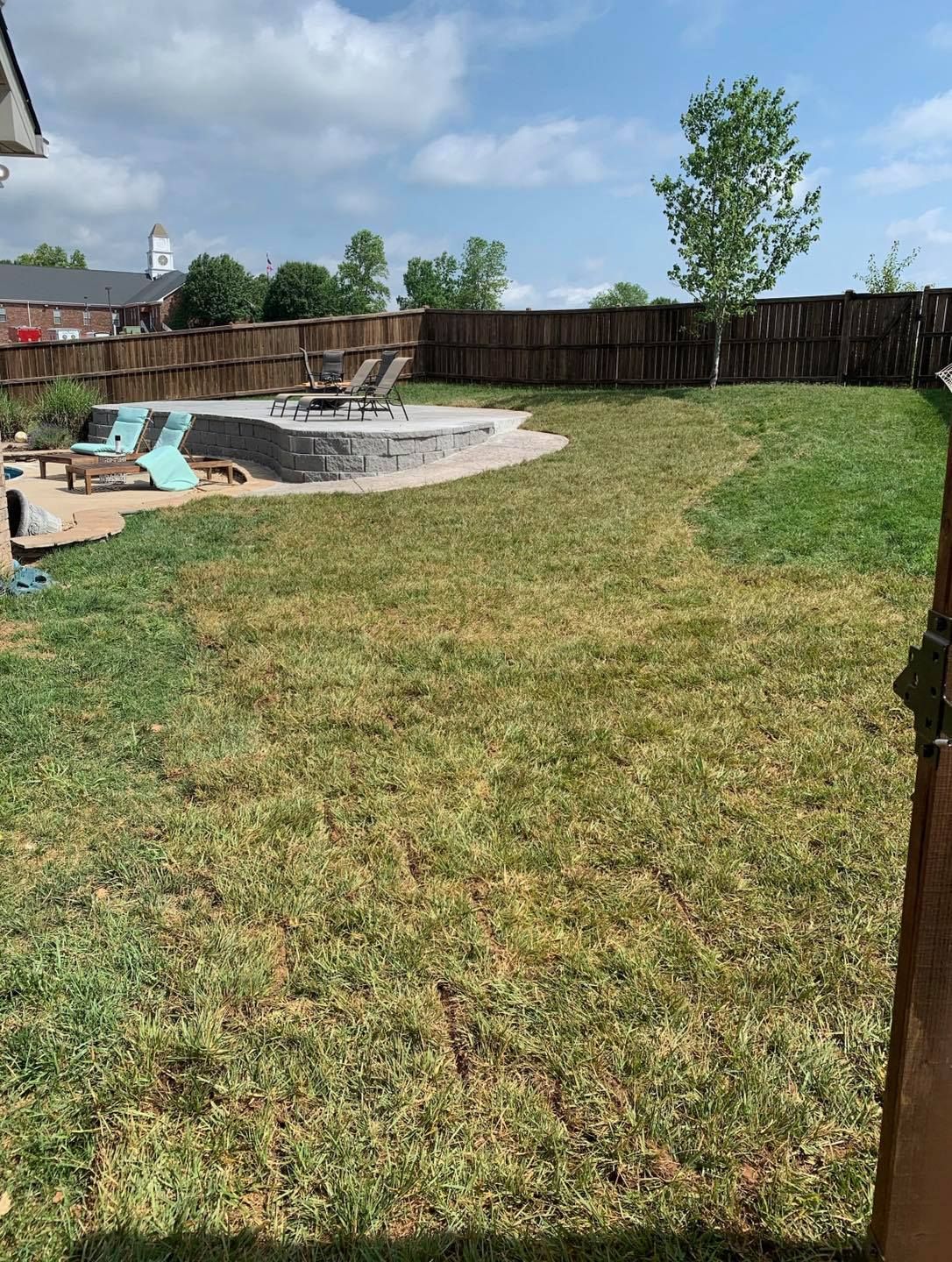 Backyard with a fire pit, chairs, and a fence, with a mix of green and dry grass under a blue sky.