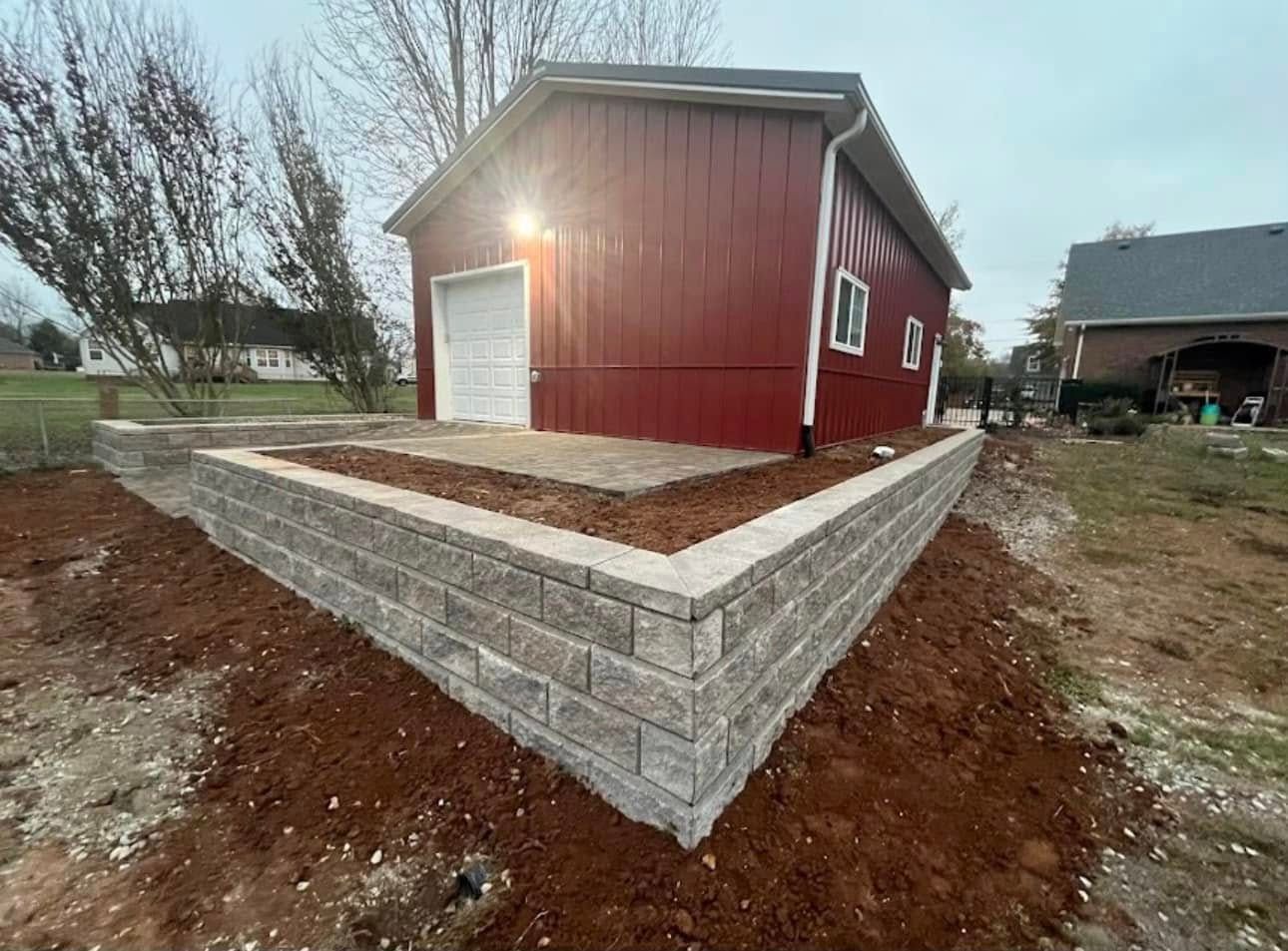 Red metal garage on a raised platform with a gray brick retaining wall. Mulch covers the platform and surrounding hillside.
