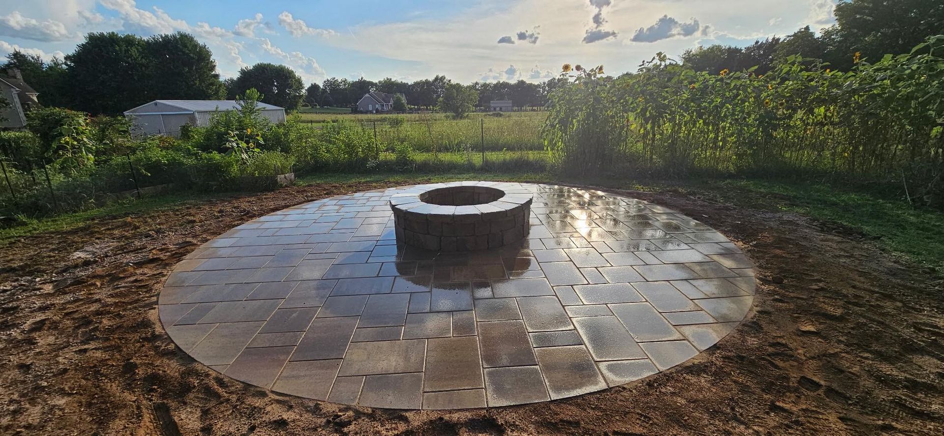 A circular paved area surrounds a stone fire pit in a grassy field. The sky is blue with clouds.