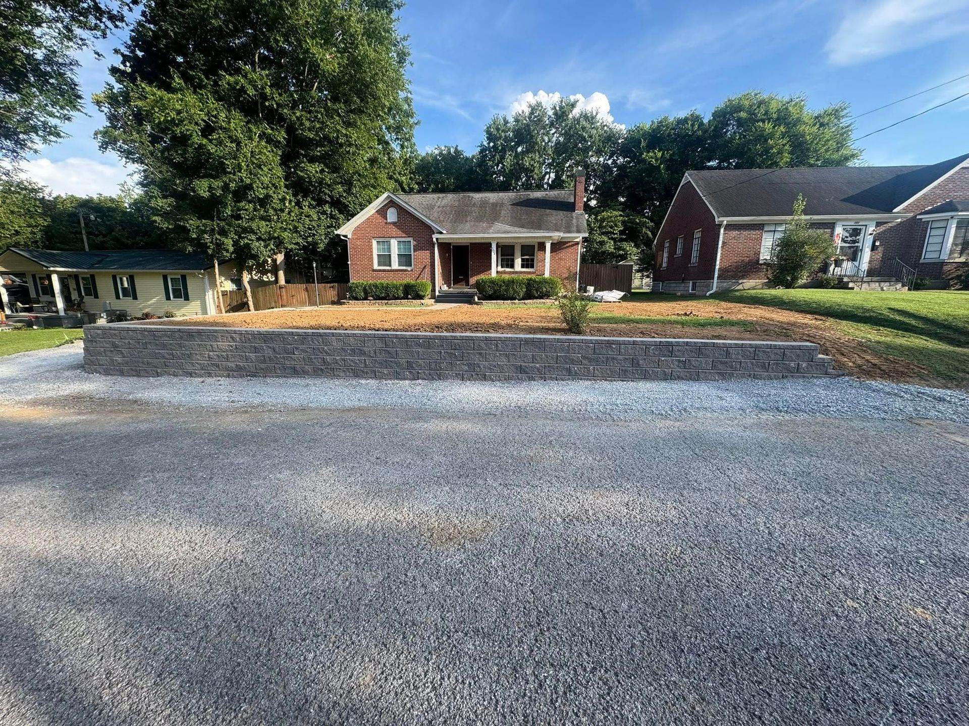 Gravel driveway in front of a brick retaining wall and small house. The house has a brown roof and a small front porch.
