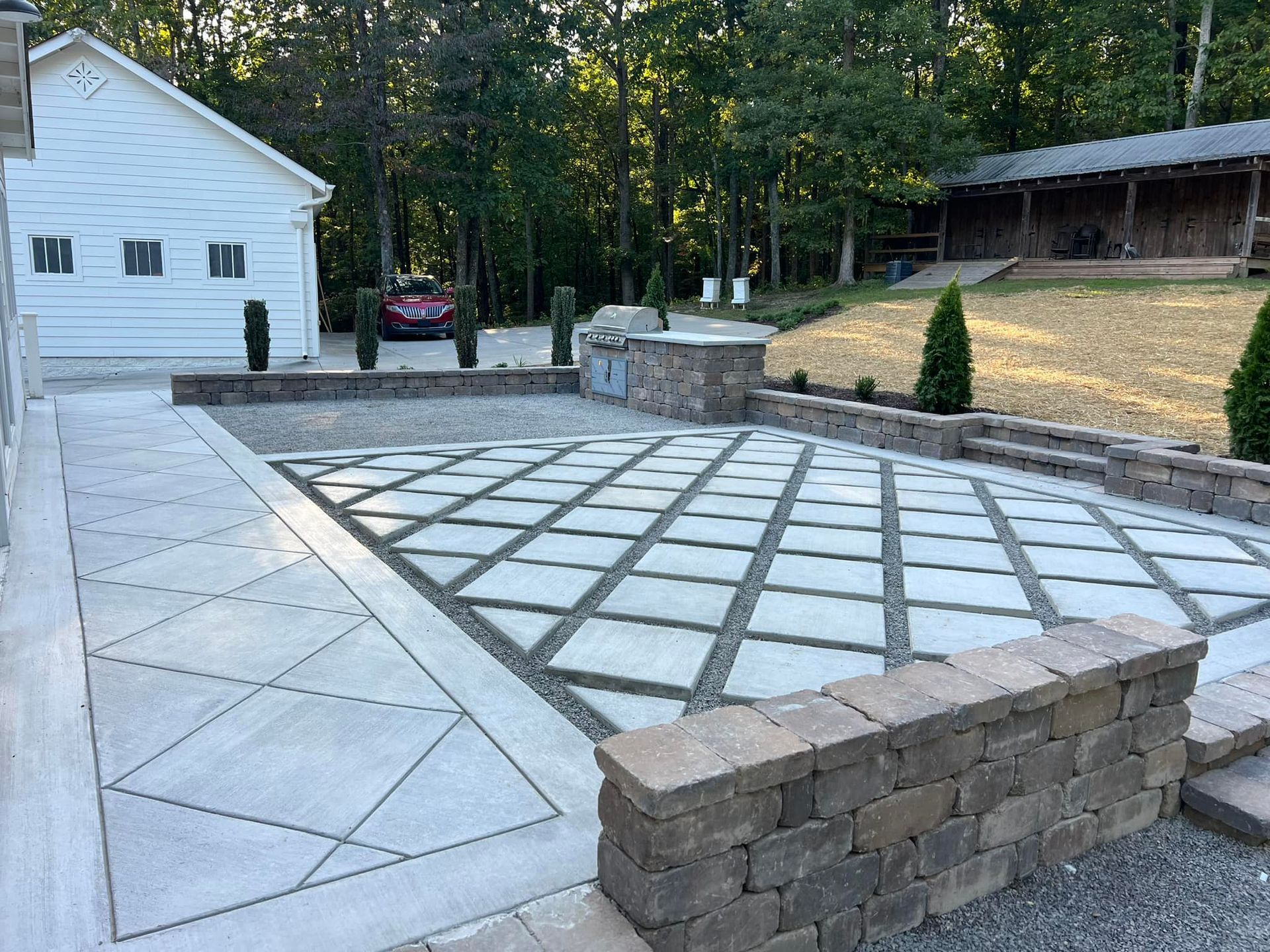 A paved outdoor patio with a stone retaining wall, adjacent to a white building and a forested area.